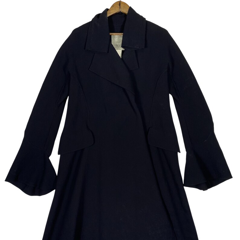 May include: A black coat with a unique design. The coat features a wide collar, long sleeves with flared cuffs, and a tailored waist. The coat is hanging on a wooden hanger. The overall style is elegant and sophisticated.
