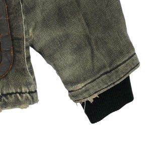 May include: Close-up of a distressed denim jacket sleeve. The jacket is a faded olive green with a black ribbed cuff. The fabric shows signs of wear and tear, with frayed edges.