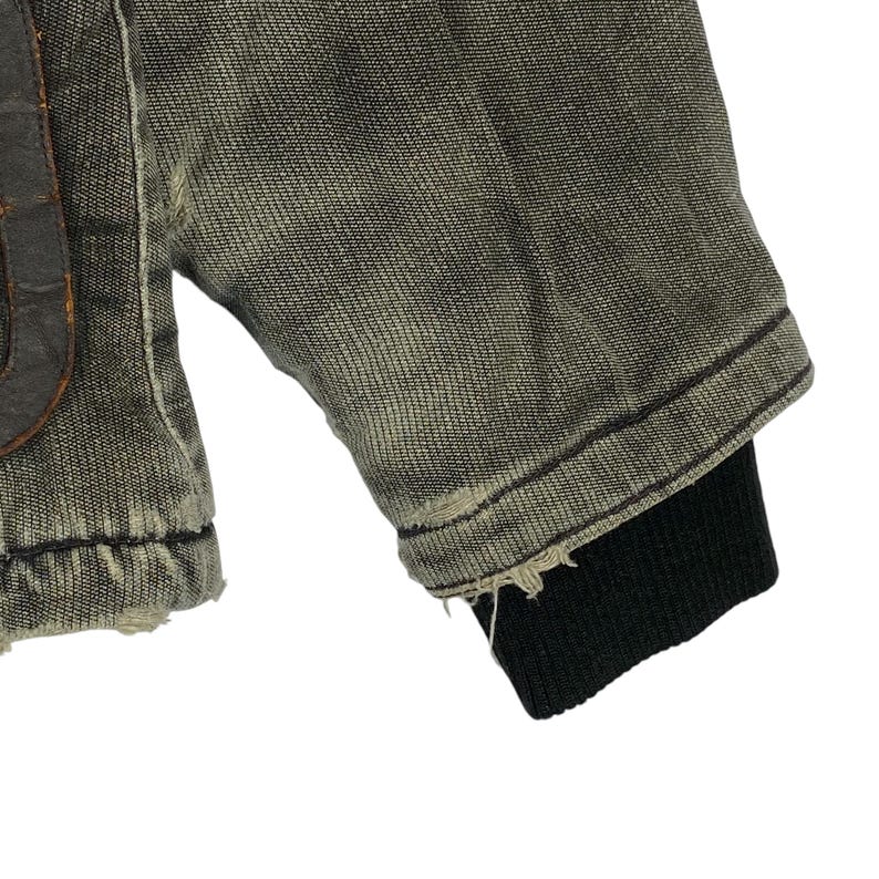May include: Close-up of a distressed denim jacket sleeve. The jacket is a faded olive green with a black ribbed cuff. The fabric shows signs of wear and tear, with frayed edges.