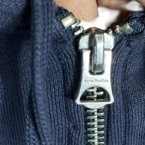 May include: Close-up of a navy blue hoodie zipper with a silver metal pull. The zipper pull is engraved with the text "Acne Studios". The zipper is fully closed, showing the teeth and the fabric of the hoodie.