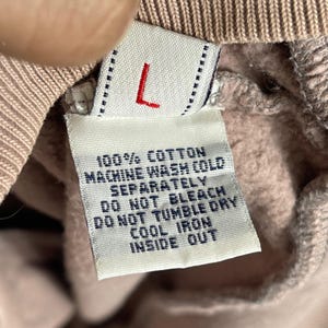 Puede incluir: Primer plano de una etiqueta de ropa con la letra "L" en rojo. La etiqueta incluye instrucciones de cuidado: 100% algod&oacute;n, lavar a m&aacute;quina en fr&iacute;o por separado, no usar lej&iacute;a, no usar secadora, planchar en fr&iacute;o del rev&eacute;s.