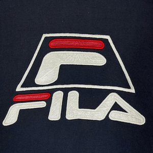 Puede incluir: Primer plano de una prenda azul marino con el logotipo de FILA. El logotipo es un dise&ntilde;o estilizado en blanco con detalles en rojo, y la palabra "FILA" est&aacute; bordada en blanco debajo. El logotipo est&aacute; centrado en la prenda.