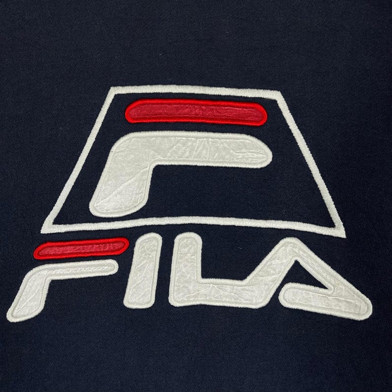Puede incluir: Primer plano de una prenda azul marino con el logotipo de FILA. El logotipo es un dise&ntilde;o estilizado en blanco con detalles en rojo, y la palabra "FILA" est&aacute; bordada en blanco debajo. El logotipo est&aacute; centrado en la prenda.