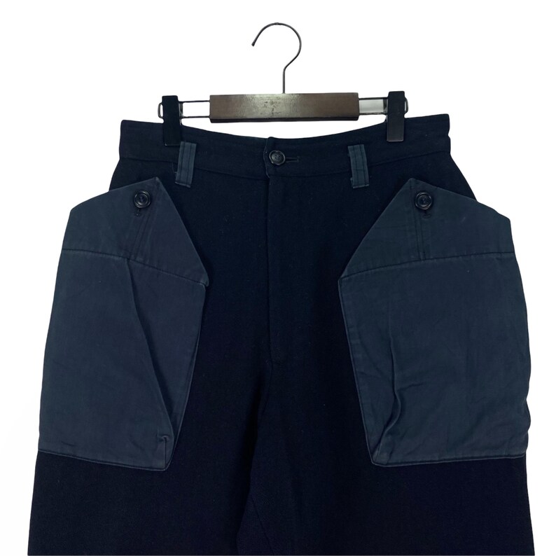 May include: Dark navy blue pants with large, contrasting navy blue pockets. The pants are hanging on a wooden hanger. The pockets have button closures. The pants are made of a sturdy material.