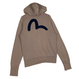 Puede incluir: Una sudadera con capucha de color beige con cord&oacute;n y mangas largas. La parte trasera de la sudadera con capucha presenta un gran s&iacute;mbolo azul marino estilizado. La sudadera tiene pu&ntilde;os y dobladillo acanalados.
