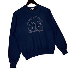 May include: A navy blue sweatshirt with white stitched text reading "sport futur" and "AC courr&egrave;ges". The sweatshirt has a crew neck and long sleeves, and is suspended from a black hanger.