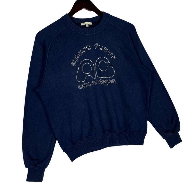 May include: A navy blue sweatshirt with white stitched text reading "sport futur" and "AC courr&egrave;ges". The sweatshirt has a crew neck and long sleeves, and is suspended from a black hanger.