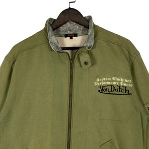 Puede incluir: Chaqueta verde oliva con cremallera y el texto "Custom Machined Performance Parts Von Dutch" en negro. La chaqueta tiene un cuello gris y un peque&ntilde;o logotipo.