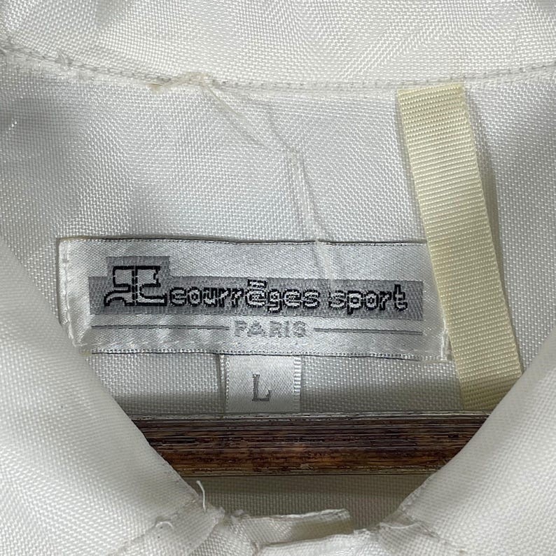 May include: White collared shirt with a tag that reads "courr&egrave;ges sport PARIS" in black text. The tag also has a logo and the size "L" printed on it. The shirt is made of a textured fabric.