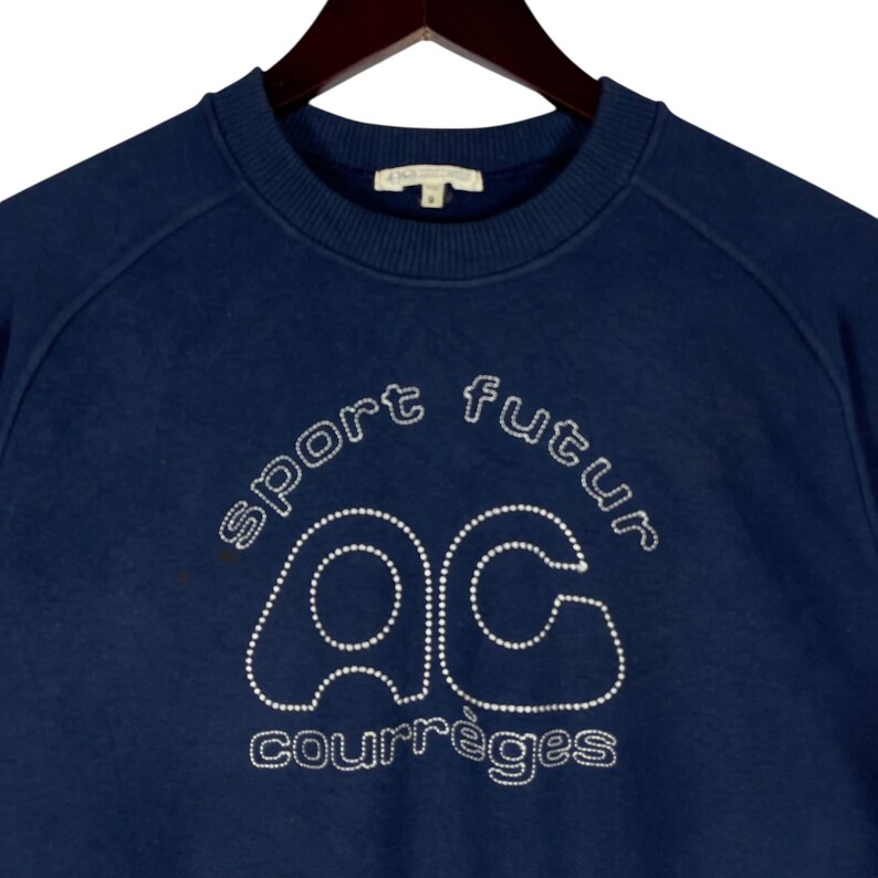 May include: Navy blue sweatshirt featuring the words "sport futur" and "courr&egrave;ges" in white, and the letters "ac" in a dotted design. The sweatshirt has a crew neck and raglan sleeves.
