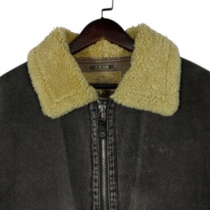 May include: Close-up of a dark brown jacket with a beige shearling collar. The jacket has a full zip closure and a small label on the inside. The jacket is hanging on a wooden hanger.