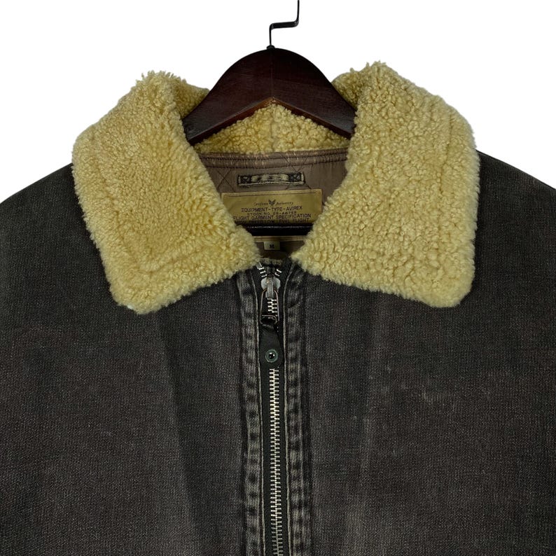 May include: Close-up of a dark brown jacket with a beige shearling collar. The jacket has a full zip closure and a small label on the inside. The jacket is hanging on a wooden hanger.