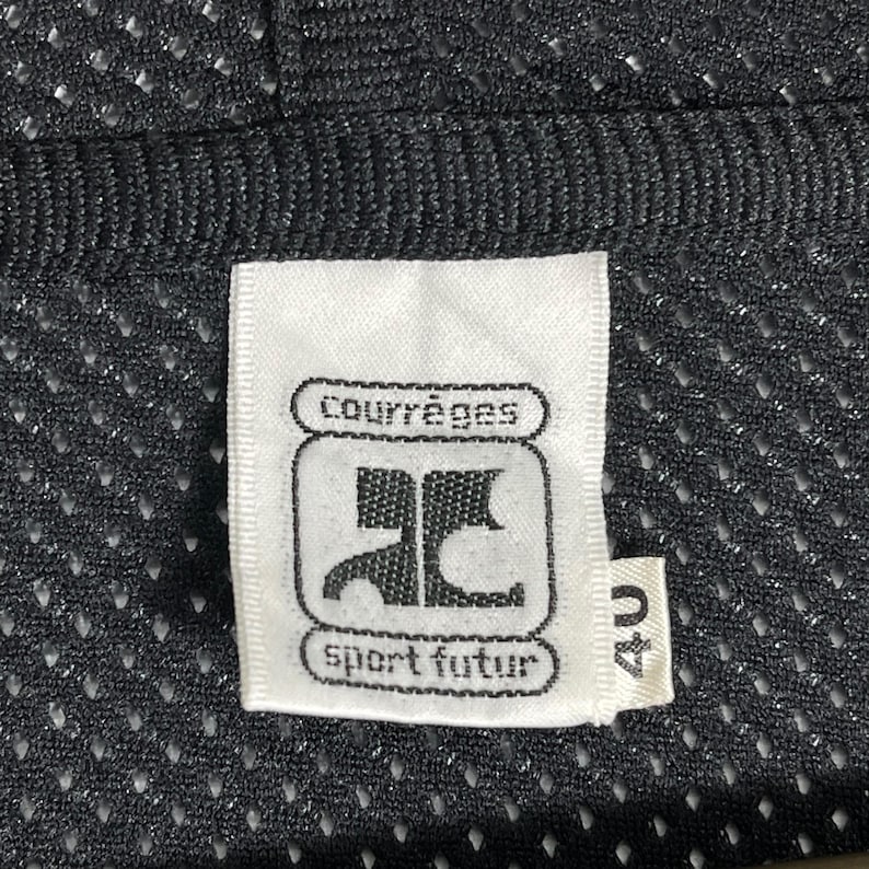 Pu&ograve; includere: Primo piano di un'etichetta bianca con il logo Courr&egrave;ges e la scritta "sport futur", su un tessuto a rete nero. L'etichetta riporta anche il numero "40".