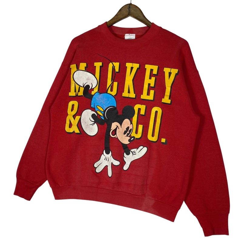May include: Red sweatshirt featuring a graphic of Mickey Mouse in a playful pose. The text "MICKEY & CO." is printed in yellow. The sweatshirt has long sleeves and a crew neck.