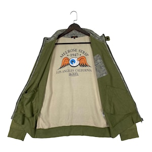 Puede incluir: Chaqueta verde oliva con cremallera y estampado de camuflaje en el cuello y los hombros. La chaqueta tiene dos bolsillos delanteros y un interior beige con un gr&aacute;fico que dice "MELROSE STRIP 1947 LOS ANGELES CALIFORNIA Von Dutch".