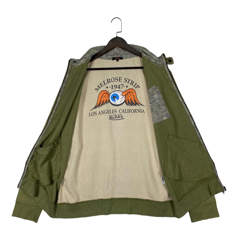 Puede incluir: Chaqueta verde oliva con cremallera y estampado de camuflaje en el cuello y los hombros. La chaqueta tiene dos bolsillos delanteros y un interior beige con un gr&aacute;fico que dice "MELROSE STRIP 1947 LOS ANGELES CALIFORNIA Von Dutch".