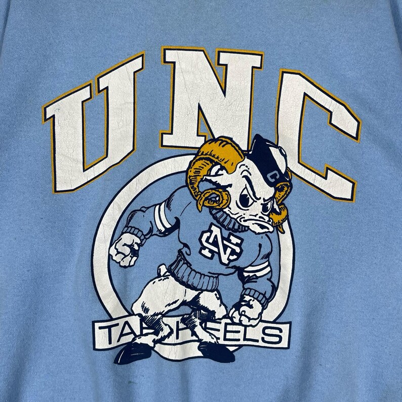 May include: A light blue sweatshirt featuring a large graphic print of a ram mascot wearing a sweater with the letters NC. Above the mascot, the letters UNC are printed in white with yellow outlines. The text TAR HEELS appears below the ram.