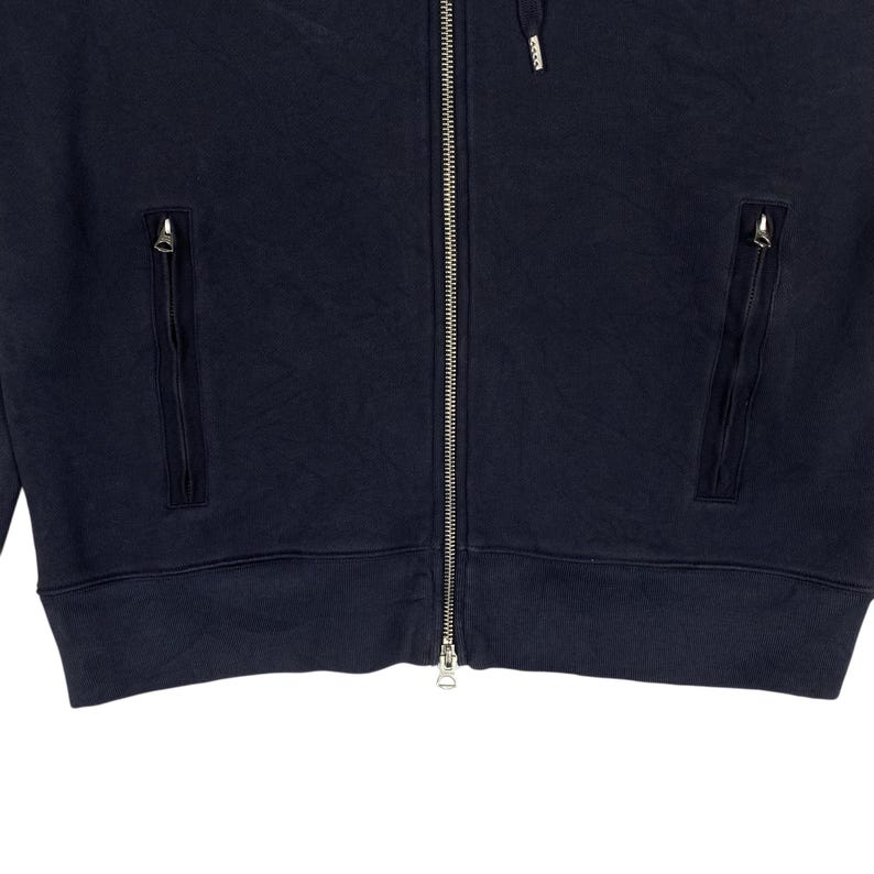 May include: Navy blue zip-up hoodie with two zippered pockets and a drawstring hood. The jacket has a full-length zipper and ribbed cuffs and hem. The fabric appears to be a soft, textured material.