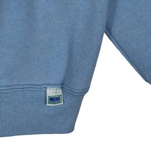 May include: A light blue sweatshirt featuring a small white rectangular fabric tag sewn onto the bottom hem. The tag displays the text "DISCUS ATHLETIC" in blue capital letters, enclosed within a thin green rectangular border on a white background.