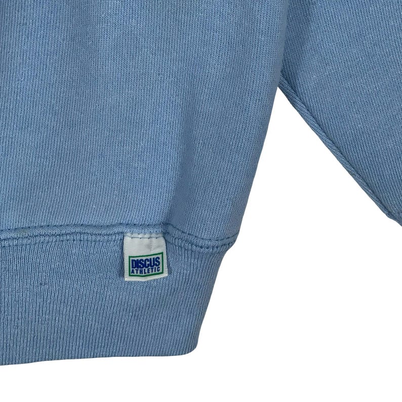 May include: A light blue sweatshirt featuring a small white rectangular fabric tag sewn onto the bottom hem. The tag displays the text "DISCUS ATHLETIC" in blue capital letters, enclosed within a thin green rectangular border on a white background.