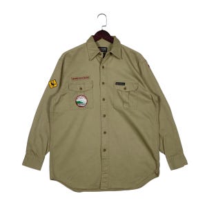 May include: A tan long-sleeve button-down shirt featuring two chest pockets, a pointed collar, and multiple embroidered patches. The patches include the text Wrangler Outdoors, a North Carolina U.S.A. emblem, and a yellow square patch.