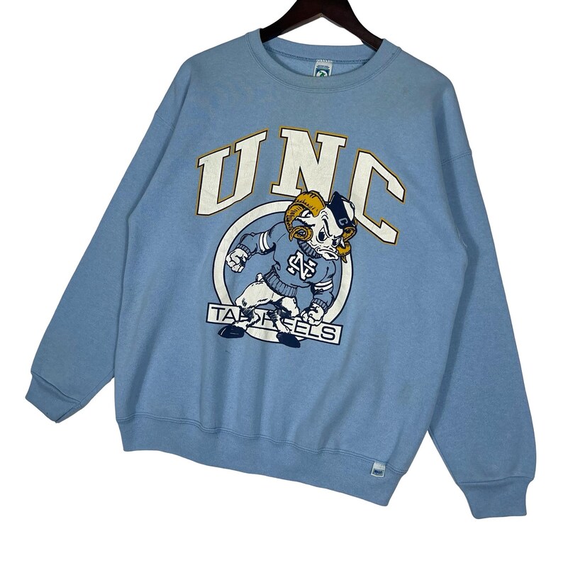 May include: A light blue crewneck sweatshirt featuring a large graphic print on the chest. The design includes the text UNC in white with gold outlines, a cartoon ram mascot wearing a blue sweater, and the text TAR HEELS in a rectangular box.