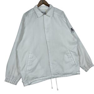 May include: A white coach jacket with a collared neckline, button-up front, and two front pockets. The jacket has a drawstring hem and a small logo on the upper left sleeve. The jacket is hanging on a wooden hanger.