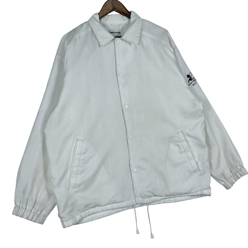 May include: A white coach jacket with a collared neckline, button-up front, and two front pockets. The jacket has a drawstring hem and a small logo on the upper left sleeve. The jacket is hanging on a wooden hanger.
