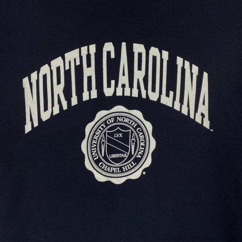 May include: Navy blue garment featuring the arched text "NORTH CAROLINA" above a circular emblem. The emblem reads "UNIVERSITY OF NORTH CAROLINA CHAPEL HILL" with the Latin words "LVX" and "LIBERTAS" in the center. The design is in white.