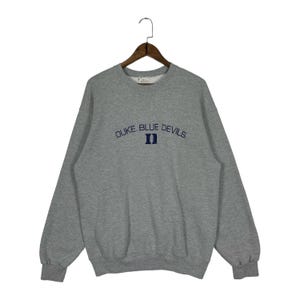 Może przedstawiać: Szara melanżowa bluza z okrągłym dekoltem, z napisem DUKE BLUE DEVILS ciemnoniebieskimi literami wygiętymi nad ciemnoniebieskim logo Duke D na piersi. Sweter z długim rękawem ma ściągacze przy mankietach i u dołu, zawieszony na drewnianym wieszaku.