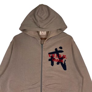 Puede incluir: Una sudadera con capucha y cremallera de color beige con capucha y cremallera frontal. La sudadera con capucha presenta un dise&ntilde;o bordado en azul marino y rojo en el pecho que dice "E.V.I.S.U. TRACK".