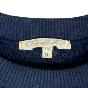 May include: Close-up of a navy blue sweatshirt with a white tag. The tag has Japanese text and the number 9. The sweatshirt is made of a soft, textured fabric.