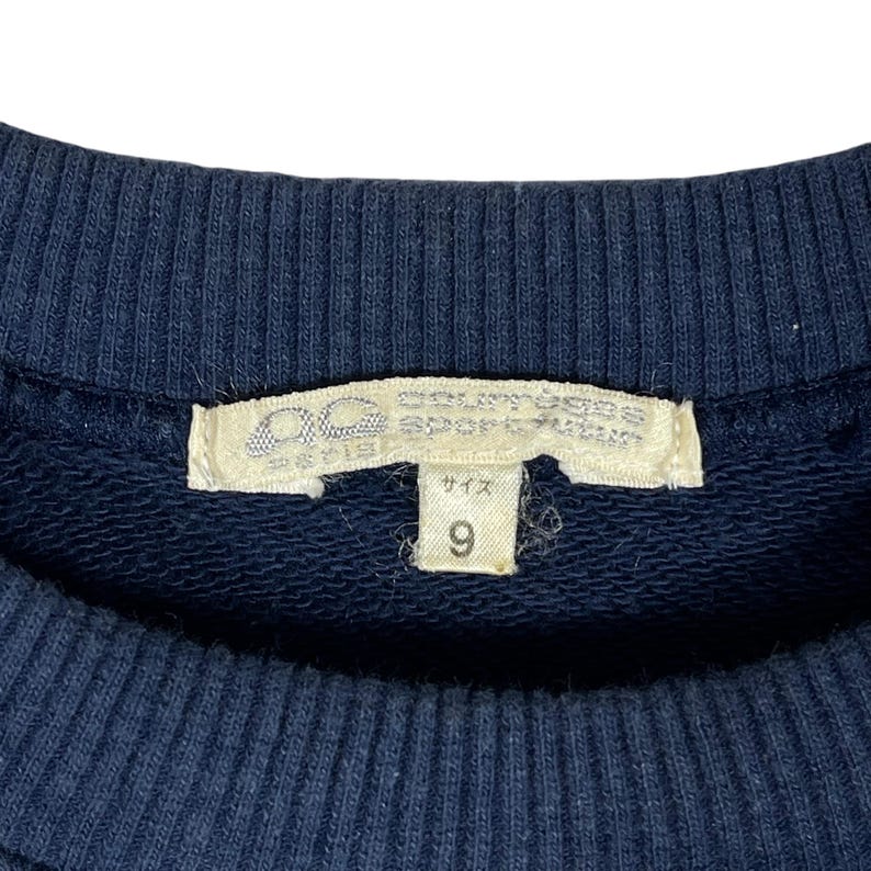 May include: Close-up of a navy blue sweatshirt with a white tag. The tag has Japanese text and the number 9. The sweatshirt is made of a soft, textured fabric.