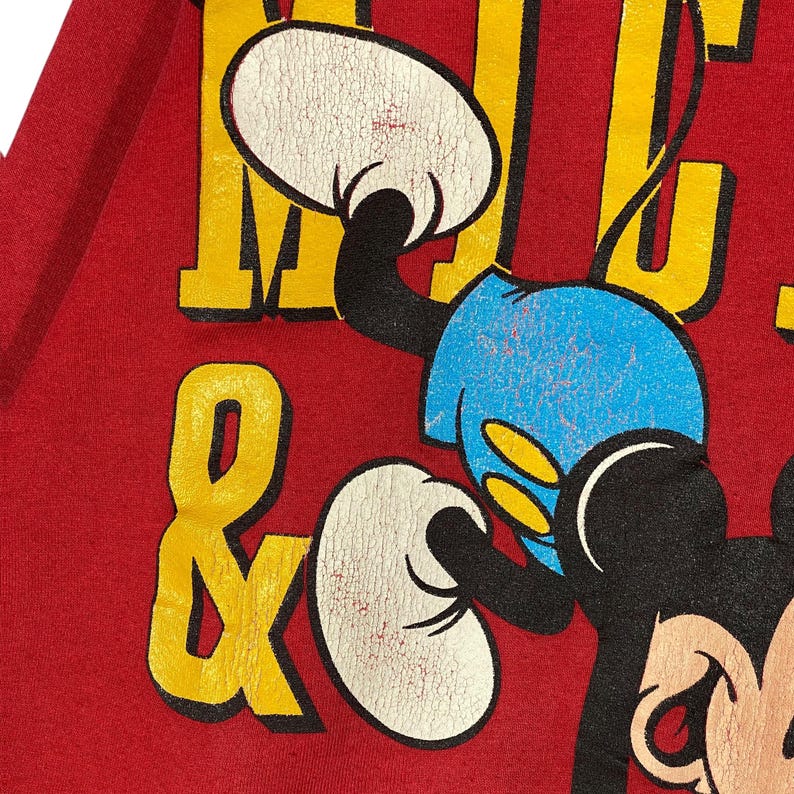 May include: Close-up of a red graphic t-shirt featuring a distressed Mickey Mouse design. The design includes yellow lettering and a blue and white Mickey Mouse illustration. The shirt has a vintage, worn look.