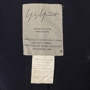 May include: Close-up of two white fabric labels with black text. The top label features a handwritten signature and the text "FABRIQUE AU JAPON MADE IN JAPAN". The bottom label includes fabric composition, care instructions, and the text "FABRIQUE AU JAPON MADE IN JAPAN".