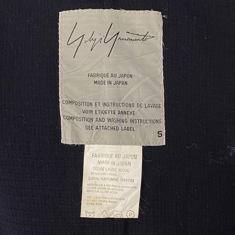 May include: Close-up of two white fabric labels with black text. The top label features a handwritten signature and the text "FABRIQUE AU JAPON MADE IN JAPAN". The bottom label includes fabric composition, care instructions, and the text "FABRIQUE AU JAPON MADE IN JAPAN".