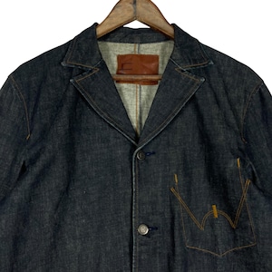 Puede incluir: Blazer vaquero de color azul oscuro con solapas y cierre de botones. Cuenta con un bolsillo en el pecho con costuras naranjas y una etiqueta de cuero marr&oacute;n. El blazer est&aacute; colgado en una percha de madera.