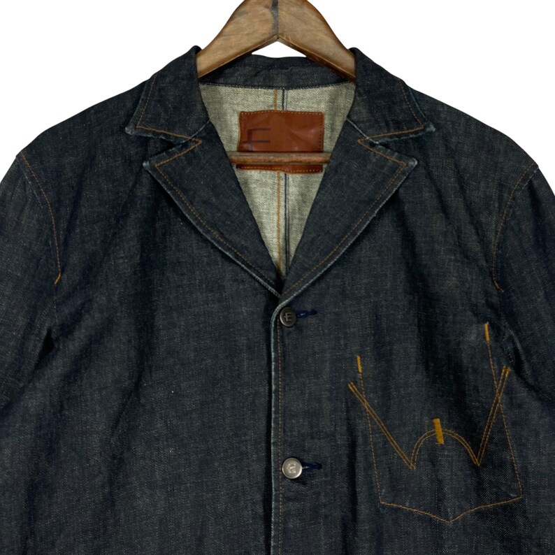 Puede incluir: Blazer vaquero de color azul oscuro con solapas y cierre de botones. Cuenta con un bolsillo en el pecho con costuras naranjas y una etiqueta de cuero marr&oacute;n. El blazer est&aacute; colgado en una percha de madera.