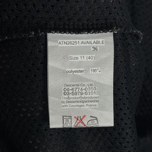 Pu&ograve; includere: Primo piano di un'etichetta con il testo "ATN26251 AVAILABLE", "36" e "Size 11 (40)". L'etichetta indica anche "polyester 100%" e include le informazioni del produttore. L'etichetta &egrave; attaccata a un tessuto a rete nero.