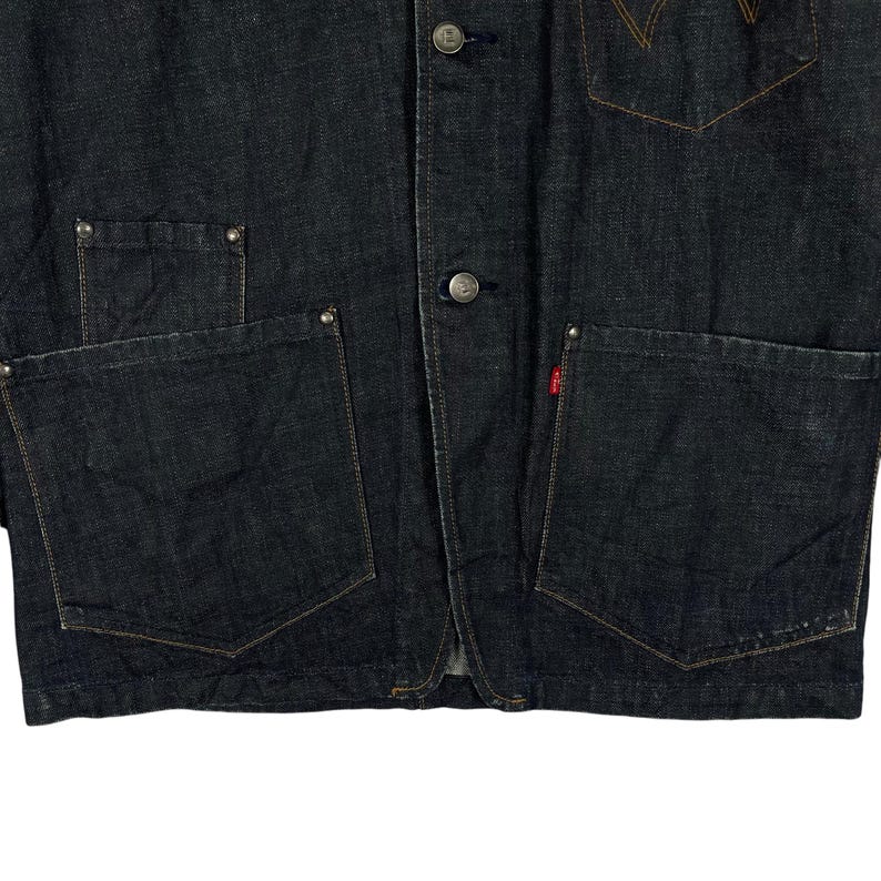 Puede incluir: Chaqueta vaquera azul oscuro con bolsillos delanteros grandes, botones y una etiqueta con el logotipo rojo. La chaqueta tiene un dise&ntilde;o cl&aacute;sico con costuras y detalles de remaches visibles.