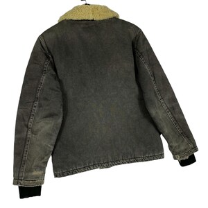 May include: A grey denim jacket with a tan sherpa collar. The jacket features black ribbed cuffs and a faded, worn look. The back view of the jacket is shown against a white background.