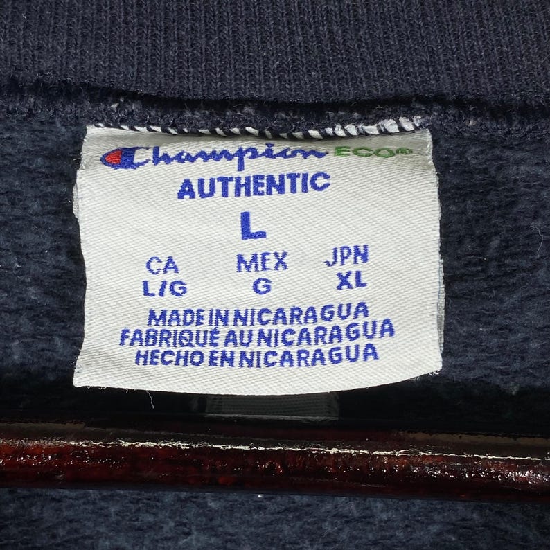 May include: Close-up of a Champion brand label on a navy blue garment. The label reads "AUTHENTIC" and includes the size "L". The label also states "MADE IN NICARAGUA" in English, French, and Spanish.