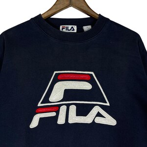 Puede incluir: Sudadera azul marino con el logotipo de FILA en blanco y rojo. El logotipo es un dise&ntilde;o estilizado con el nombre de la marca en un lugar destacado. La sudadera tiene cuello redondo y un corte cl&aacute;sico.