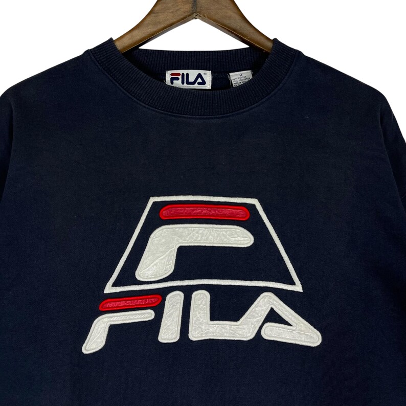 Puede incluir: Sudadera azul marino con el logotipo de FILA en blanco y rojo. El logotipo es un dise&ntilde;o estilizado con el nombre de la marca en un lugar destacado. La sudadera tiene cuello redondo y un corte cl&aacute;sico.