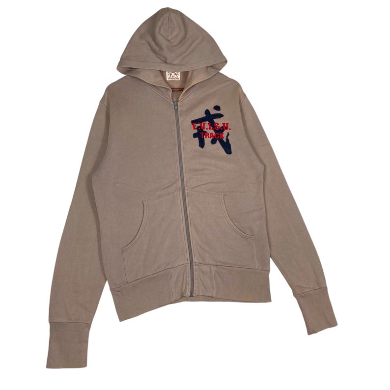 Puede incluir: Una sudadera con capucha con cremallera de color beige con capucha y bolsillos delanteros. La sudadera con capucha presenta un dise&ntilde;o bordado en azul oscuro y rojo en el pecho. Las mangas tienen pu&ntilde;os acanalados.