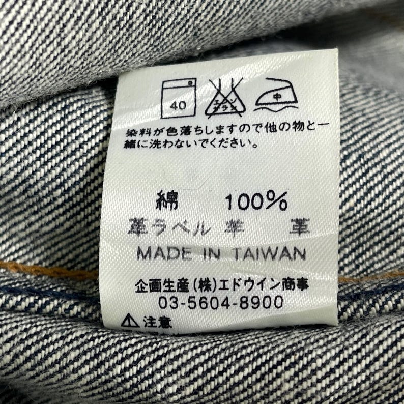 Puede incluir: Primer plano de una etiqueta de ropa blanca con texto japon&eacute;s y s&iacute;mbolos de cuidado, cosida en tejido vaquero. La etiqueta dice "100% algod&oacute;n" y "Made in Taiwan". La etiqueta tambi&eacute;n incluye instrucciones de lavado y un n&uacute;mero de tel&eacute;fono.