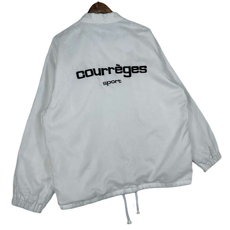 May include: White Courr&egrave;ges sport jacket with a drawstring hem. The jacket has a black logo on the back. The jacket has a collar and a zipper closure. The jacket is made of a lightweight material.