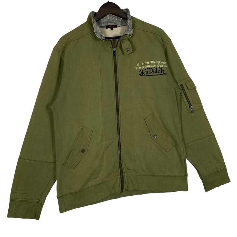 Puede incluir: Chaqueta verde oliva con cremallera y el texto "Custom Machined Performance Parts Von Dutch" bordado en el pecho. La chaqueta tiene cuello alto, dos bolsillos delanteros y un bolsillo con cremallera en la manga izquierda.