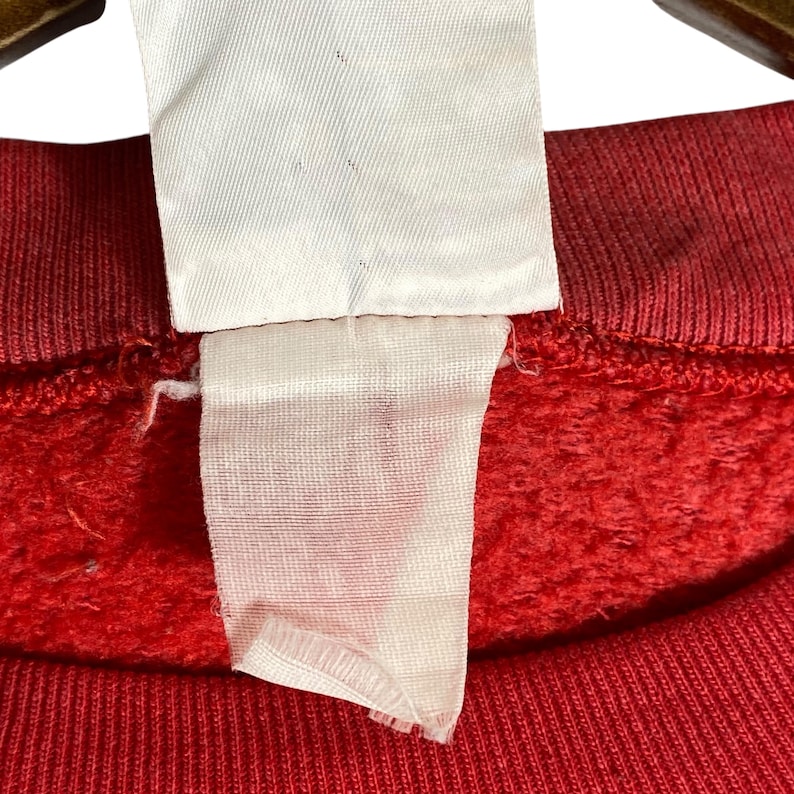 May include: Close-up of a red sweatshirt with a white tag attached. The tag is rectangular and appears to be made of fabric. The sweatshirt has a textured, fleecy appearance.