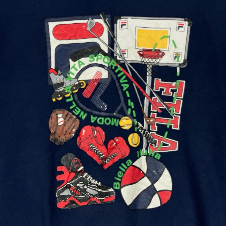 May include: A dark blue t-shirt featuring a graphic print of various sports equipment including a basketball hoop, a baseball glove, red boxing gloves, a tennis racket, golf clubs, and a sneaker. The text reads FILA, MODA NELLA VITA SPORTIVA, and Biella.